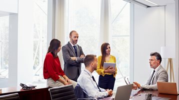 Groupe de cinq professionnels travaillant en groupe dans une salle de conférence. Trois personnes debout près de la fenêtre et deux personnes assises sur des chaises. Papiers et ordinateur portable sur la table de la salle de réunion. 