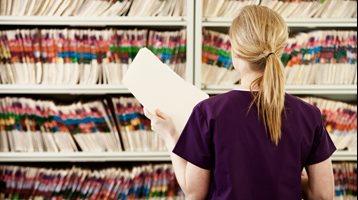 Woman in purple scrubs standing in front of two open filing cabinets with rows of paper records, holding one file in her hand. 