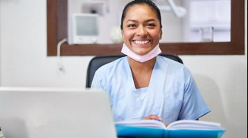 Dentiste souriante assise à son bureau devant un ordinateur portable et un carnet de notes. 